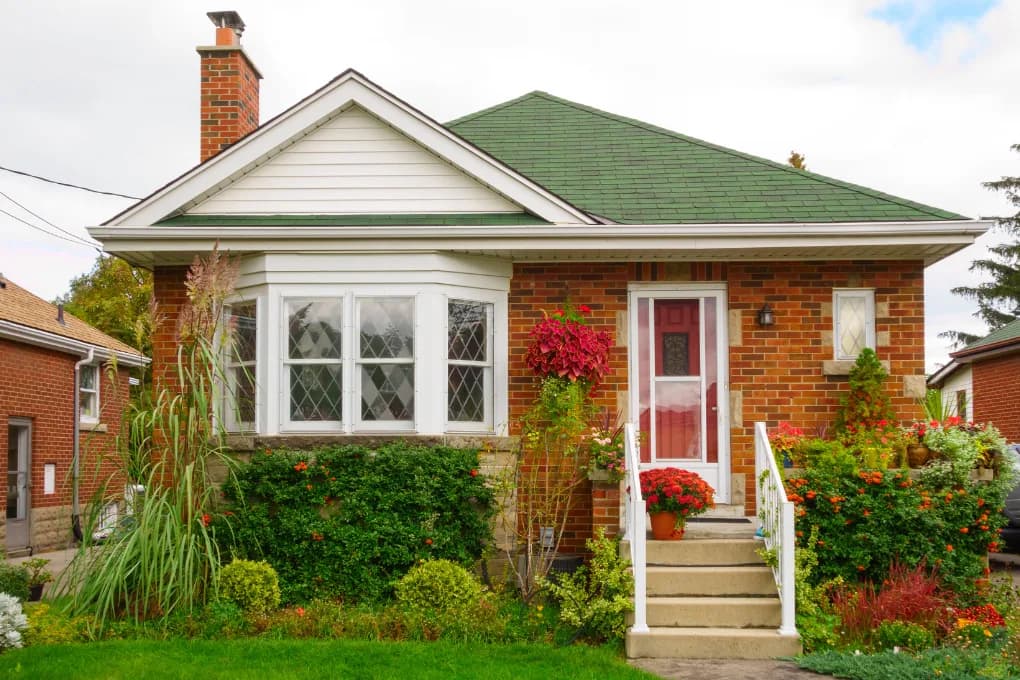 Street view of a small house with a large front window and flowers in the front yard.