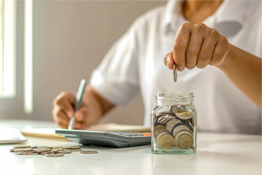 Person calculating expenses using a notepad and a calculator with one hand, while the other hand drops a coin into a jar full of coins.