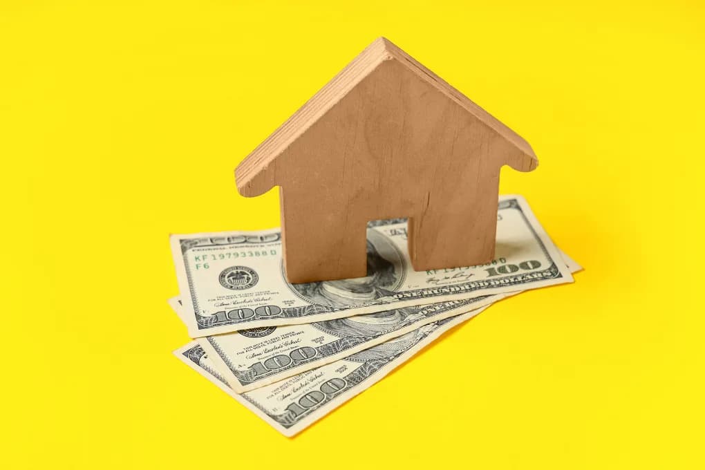 Toy house sitting on top of cash with a yellow background.